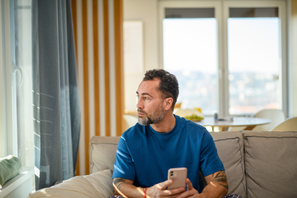 Handsome man sitting on sofa, holding smartphone in hand and looking out of window.