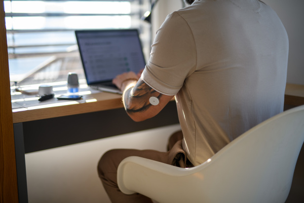 Diabetic man with CGM sensor on arm sitting at table and working on laptop.