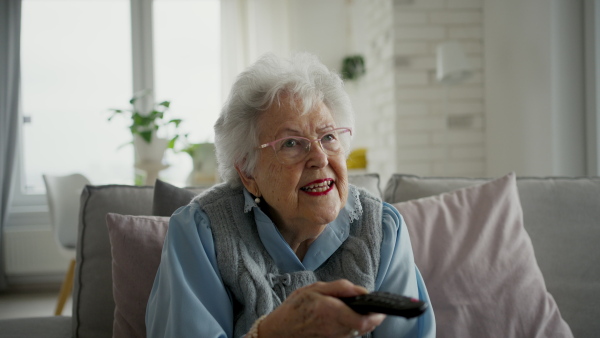 Adorable senior woman watching television, holding TV remote controller.