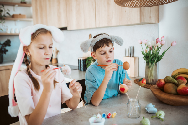 Girl and boy paiting eggs for easter, wearing bunny ears. Siblings decorate egshells for easter egg decorating.