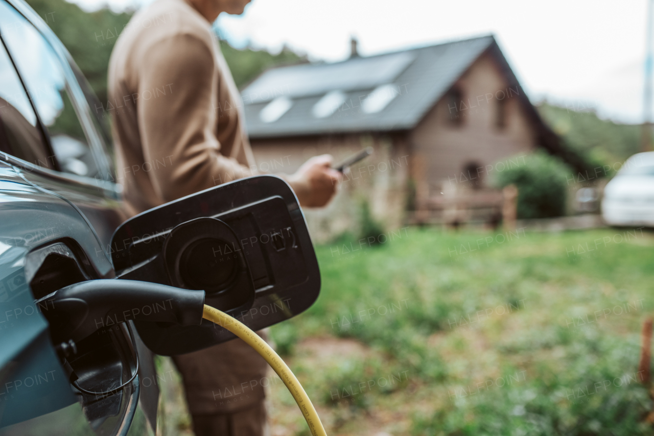 Man charging electric car, checking progress in app. House with solar panel system on roof behind him.