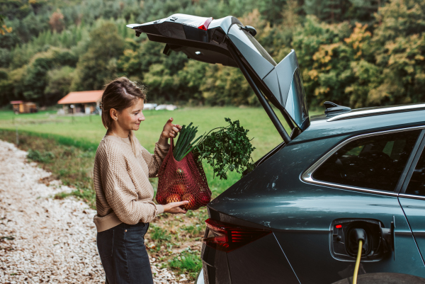 Side view of woman taking out groceries from trunk of charging electric car.