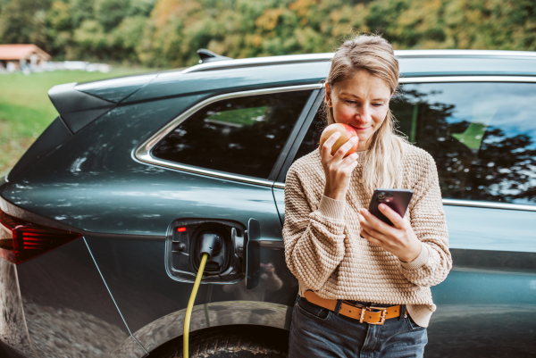 Woman charging electric car, checking progress in app while eating apple.