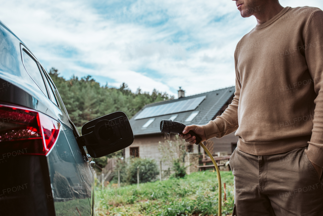 Close up of man charging electric car, plugging charger in charging port. House with solar panels behind him.