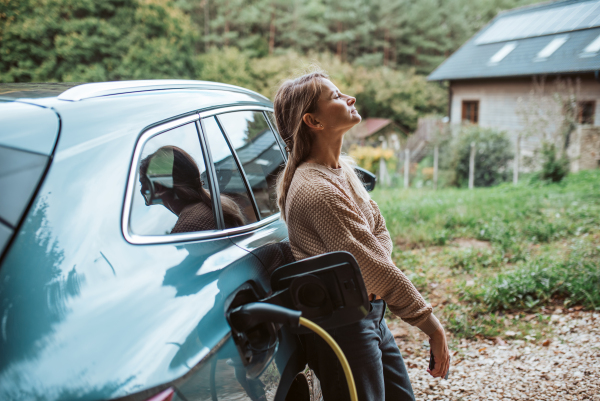 Woman charging electric car, leaning against vehicle with charger in charging port. House with solar panels on roof behind.