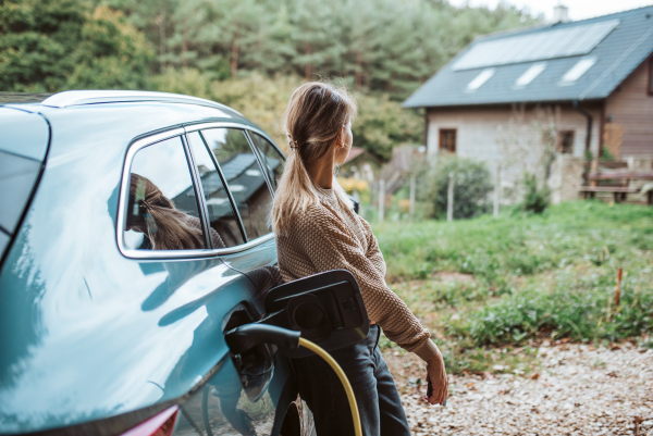 Woman charging electric car, leaning against vehicle with charger in charging port. House with solar panels on roof behind.