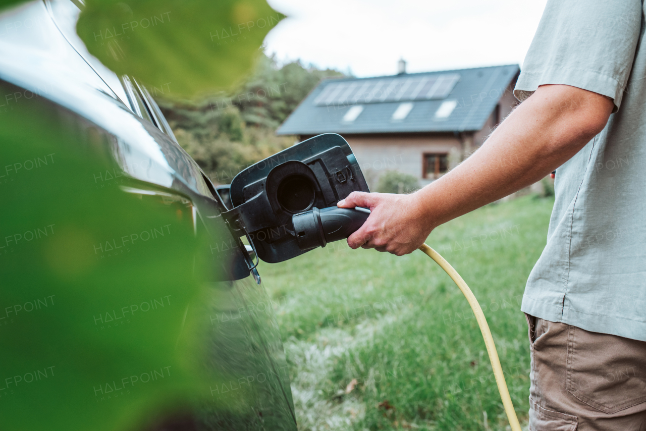 Close up of man charging electric car, plugging charger in charging port. House with solar panels behind him.