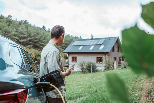 Man charging electric car, checking progress in app. House with solar panel system on roof behind him.