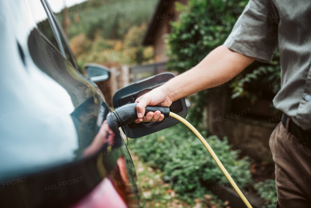 Close up of man charging electric car, plugging charger in charging port. House with solar panels behind him.