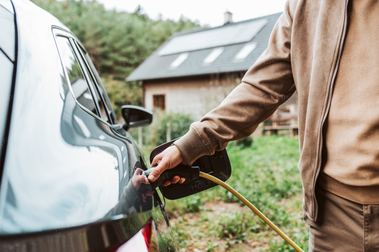 Close up of man charging electric car, plugging charger in charging port. House with solar panels behind him.