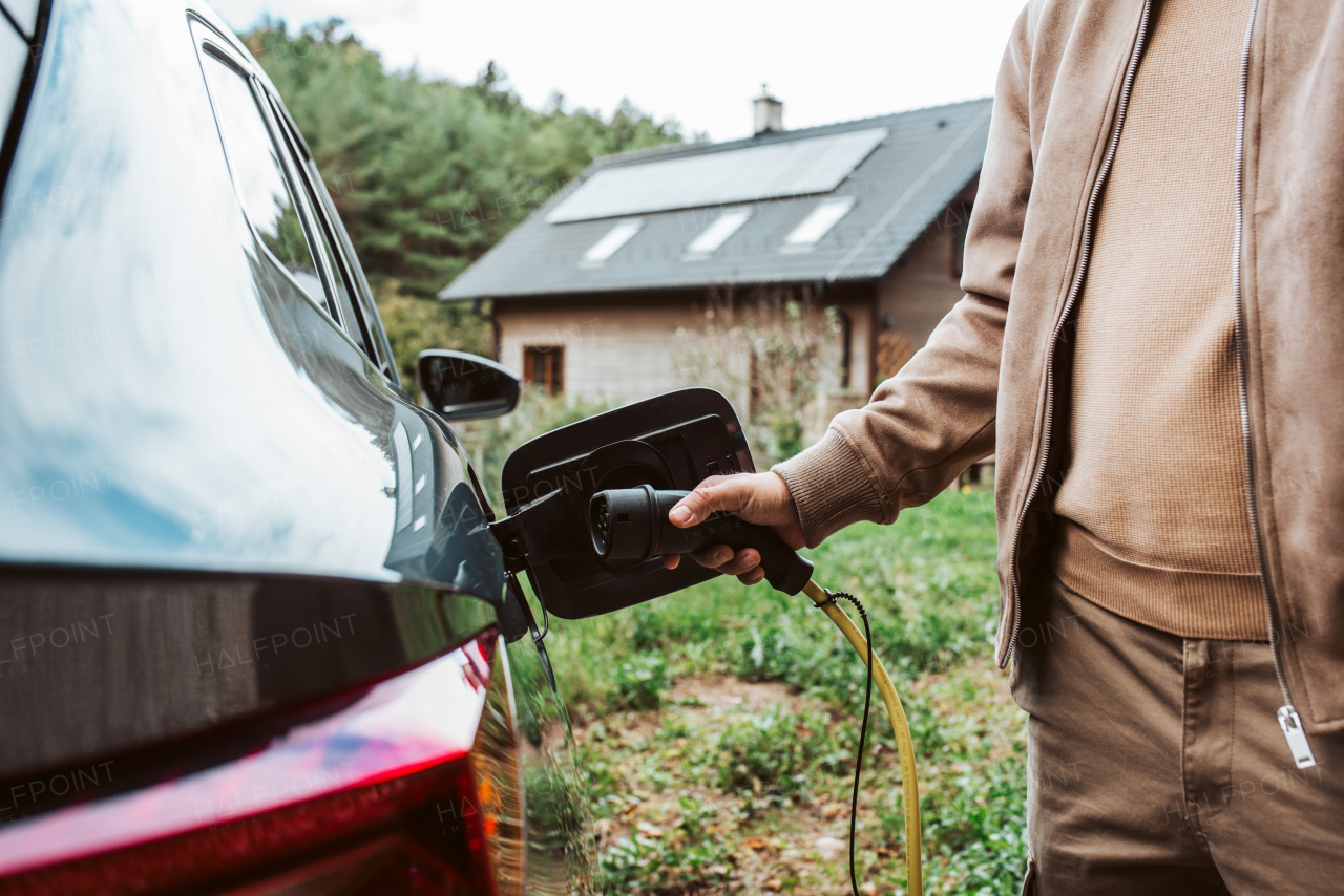 Close up of man charging electric car, plugging charger in charging port. House with solar panels behind him.