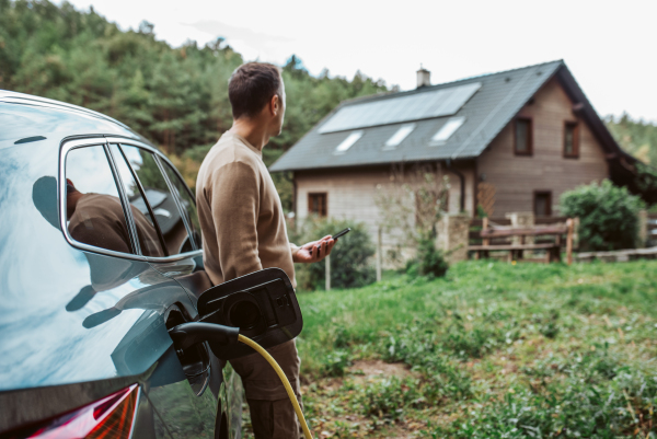 Man charging electric car, checking progress in app. House with solar panel system on roof behind him.