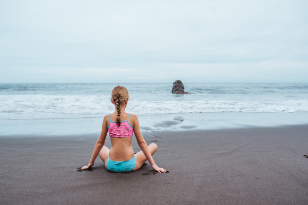 Young girl sitting on sandy beach in a swimsuit, gazing at the ocean waves.