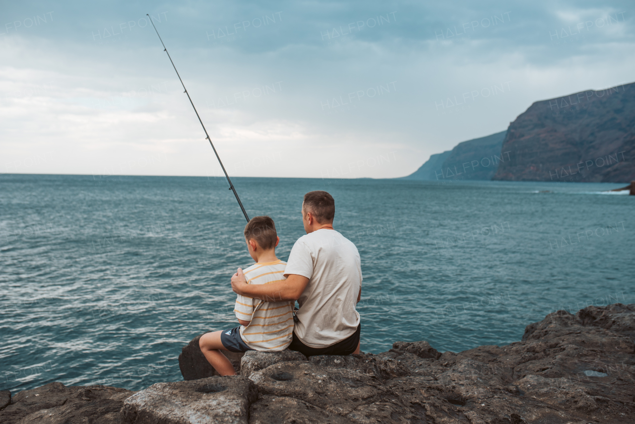 Rear view of father and young son fishing in the sea. Sitting on rock holding fish rod.