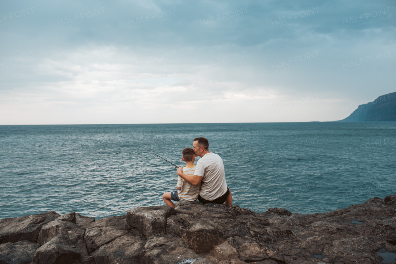 Rear view of father and young son fishing in the sea. Sitting on rock holding fish rod.