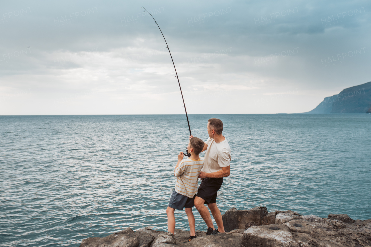 Rear view of father and young son fishing in the sea. Standing on rock holding fish rod.