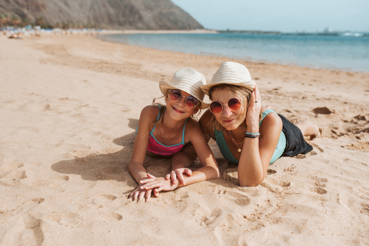 Portrait of mother and daughter lying on sandy beach, wearing hats and sunglasses.