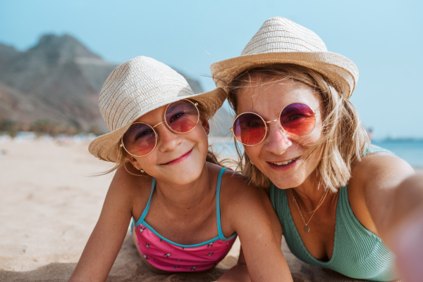 Portrait of mother and daughter on sandy beach, wearing hats and sunglasses.