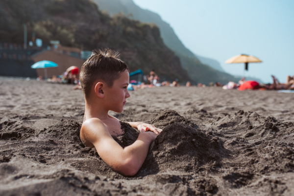 Side view of boy buried in sand on the beach, having fun during a family vacation.