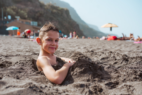 Side view of boy buried in sand on the beach, having fun during a family vacation.