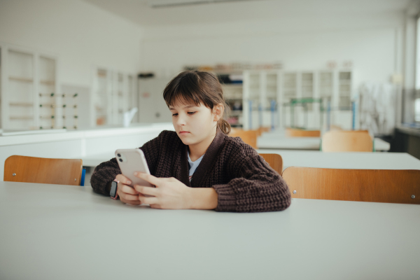 Young schoolgirl sitting in a science lab with a phone in her hand, waiting for classmates. Cyberbullying at school.