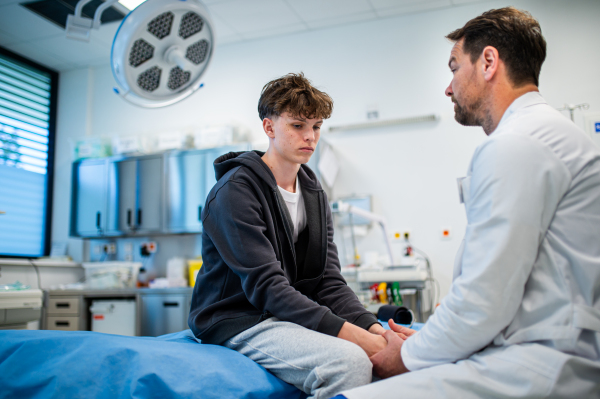 Depressed teenage boy patient sitting on hospital bed, talking with caring doctor.