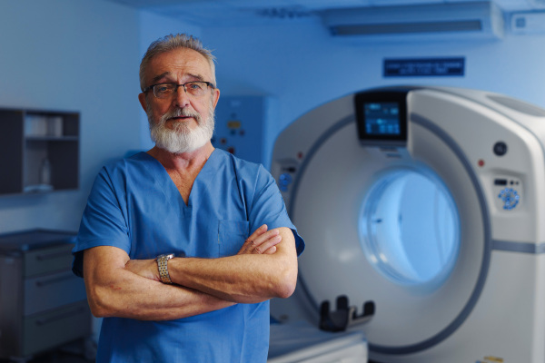 Medical specialist standing by CT scanner, looking at camera in hospital setting.