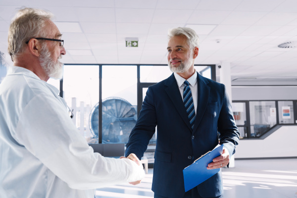 Older doctor shaking hand with hospital manager, standing in hospital hallway.