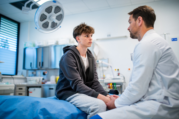 Depressed teenage boy patient sitting on hospital bed, talking with caring doctor.