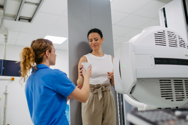 Young woman preparing for dynamic X-ray on tilting fluoroscopy wall unit, drinking contrast liquid.