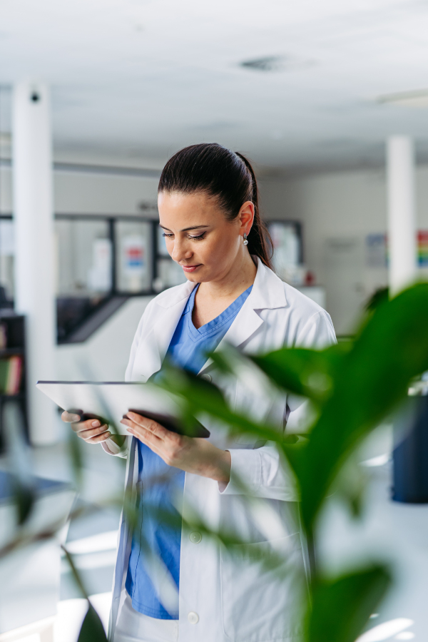 Portrait of female doctor standing in hospital hallway, reading document in tablet.