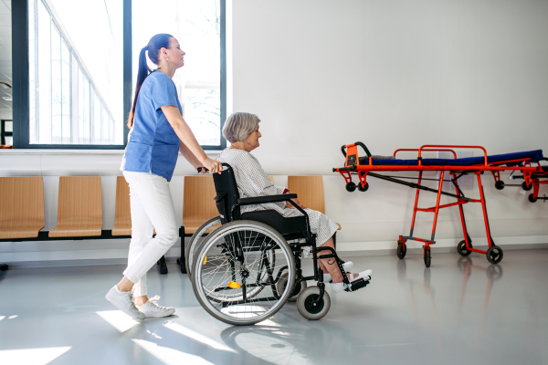 Friendly doctor pushing elderly patient in wheelchair across hospital hallway, Senior health care.
