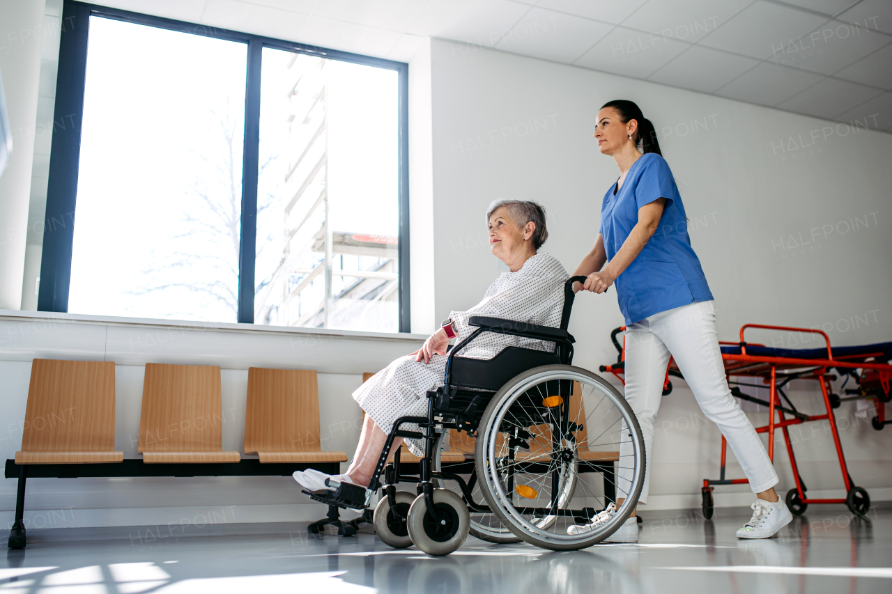 Friendly doctor pushing elderly patient in wheelchair across hospital hallway, Senior health care.
