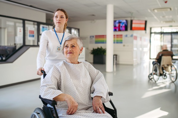 Friendly doctor pushing elderly patient in wheelchair across hospital hallway, Senior health care.