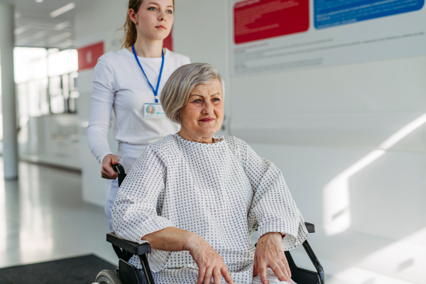 Nurse pushing elderly patient in wheelchair across hospital hallway, Senior health care.