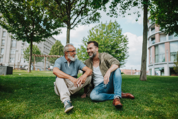 Father and adult son sitting on grass in park, sharing a joyful moment. Fathers day concept.