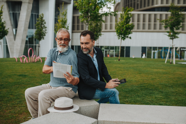 Father and adult son sitting on bench in city park, reading newspaper. Fathers day concept.