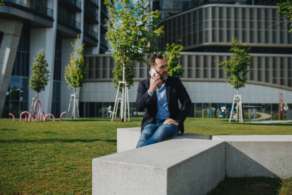 Mature businessman sitting on concrete bench in park and talking on phone