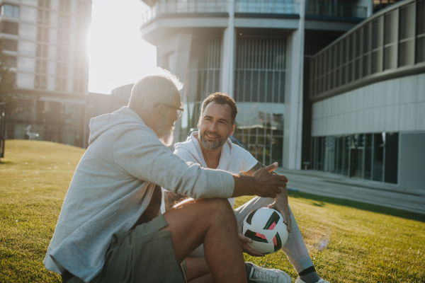 Sporty father and adult son resting after football match in park. Fathers day concept.
