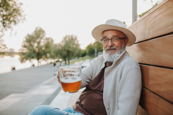 Senior man enjoying beer on sunny riverside promenade during sunset.