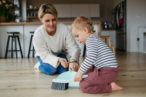 Mother teaching toddler to sweep the floor at home using dustpan and brush