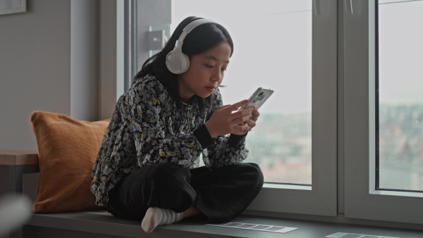 Young japanese girl with headphones scrolling her smartphone while listening to music next to window.