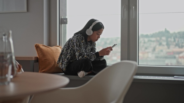 Young japanese girl with headphones scrolling her smartphone while listening to music next to window.