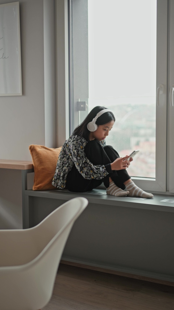 Young japanese girl with headphones scrolling her smartphone while listening to music next to window.