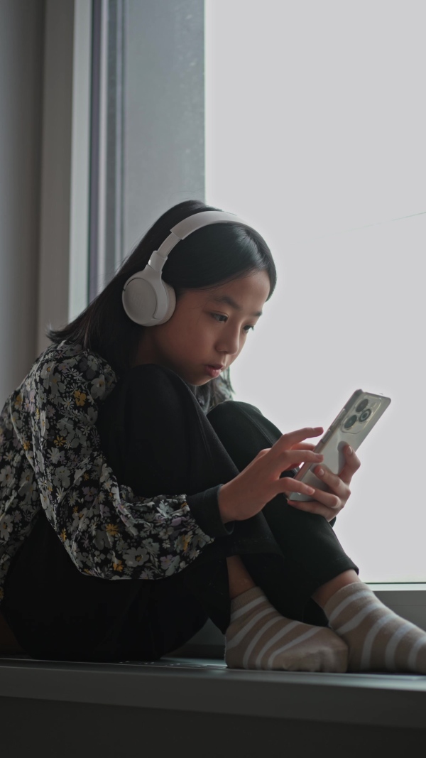 Young japanese girl with headphones scrolling her smartphone while listening to music next to window.