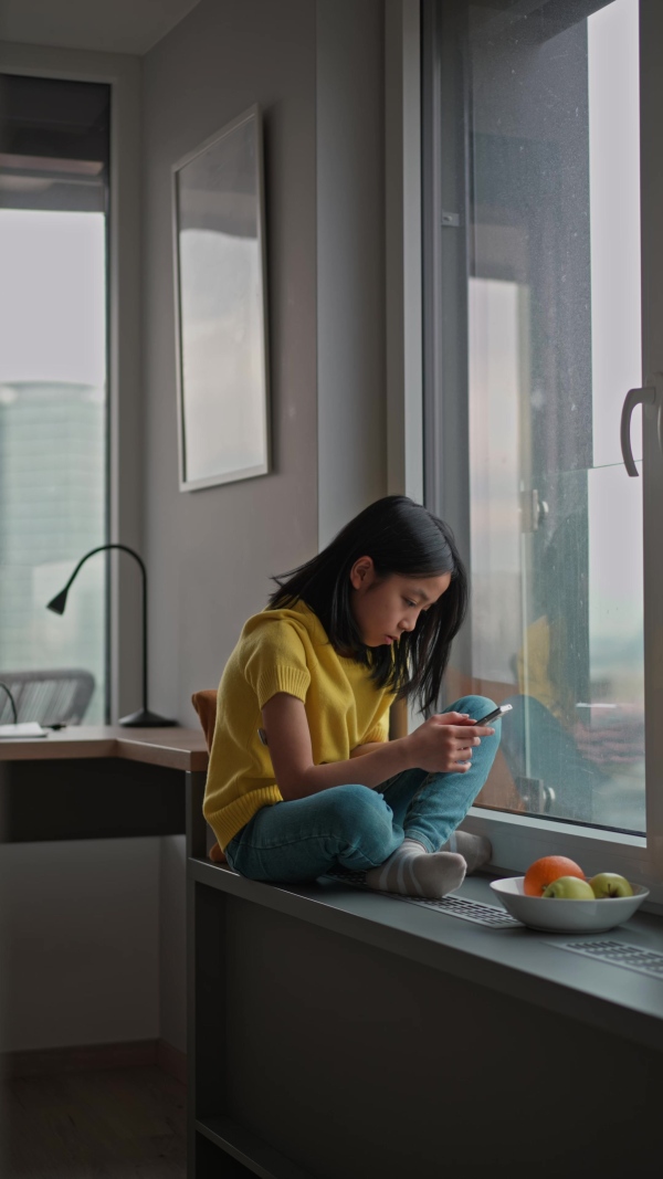 Young diabetic japanese girl with CGM sensor scrolling her smartphone next to a window.