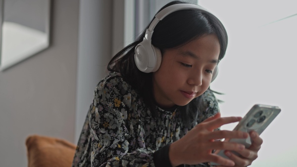 Young japanese girl with headphones scrolling her smartphone while listening to music next to window.