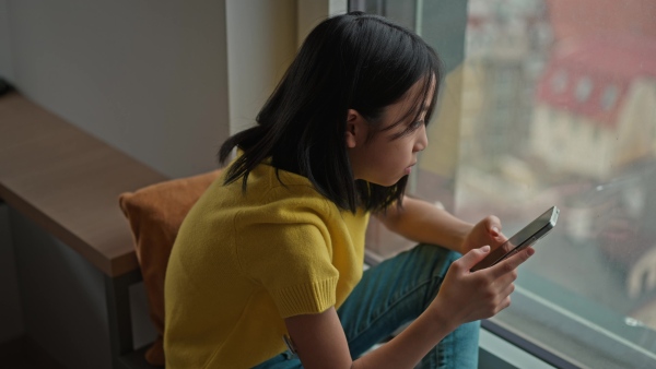 Young diabetic japanese girl with CGM sensor scrolling her smartphone next to a window.