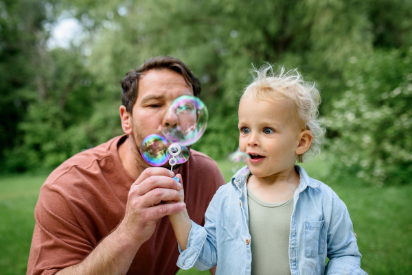 Father and son blowing soap bubbles in park. Toddler watching bubbles with amazement.