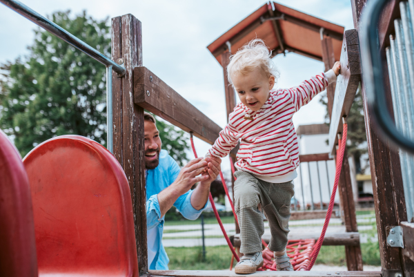 Toddler crossing playground obstacle with dad's support, weekend activity.